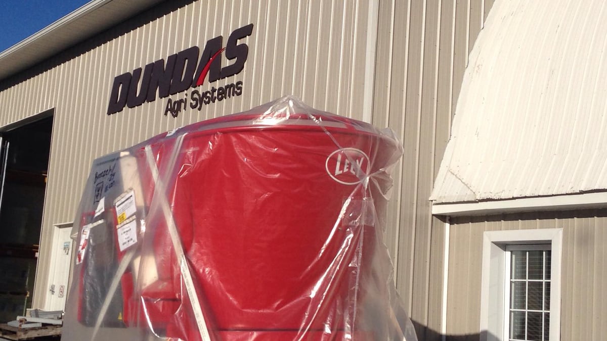 Large red machinery covered with clear plastic wrap, placed outdoors in front of a building labeled ‘DUNDAS Agri Systems,’ showcasing agricultural equipment.