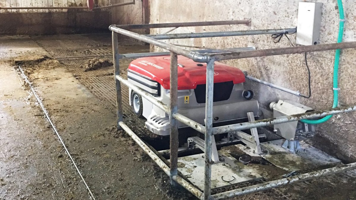 A robotic device, possibly an automated cleaning machine, located in a barn-like setting with a concrete floor. The machine is red and white and housed within a metal frame structure, which might be part of its docking or charging station.