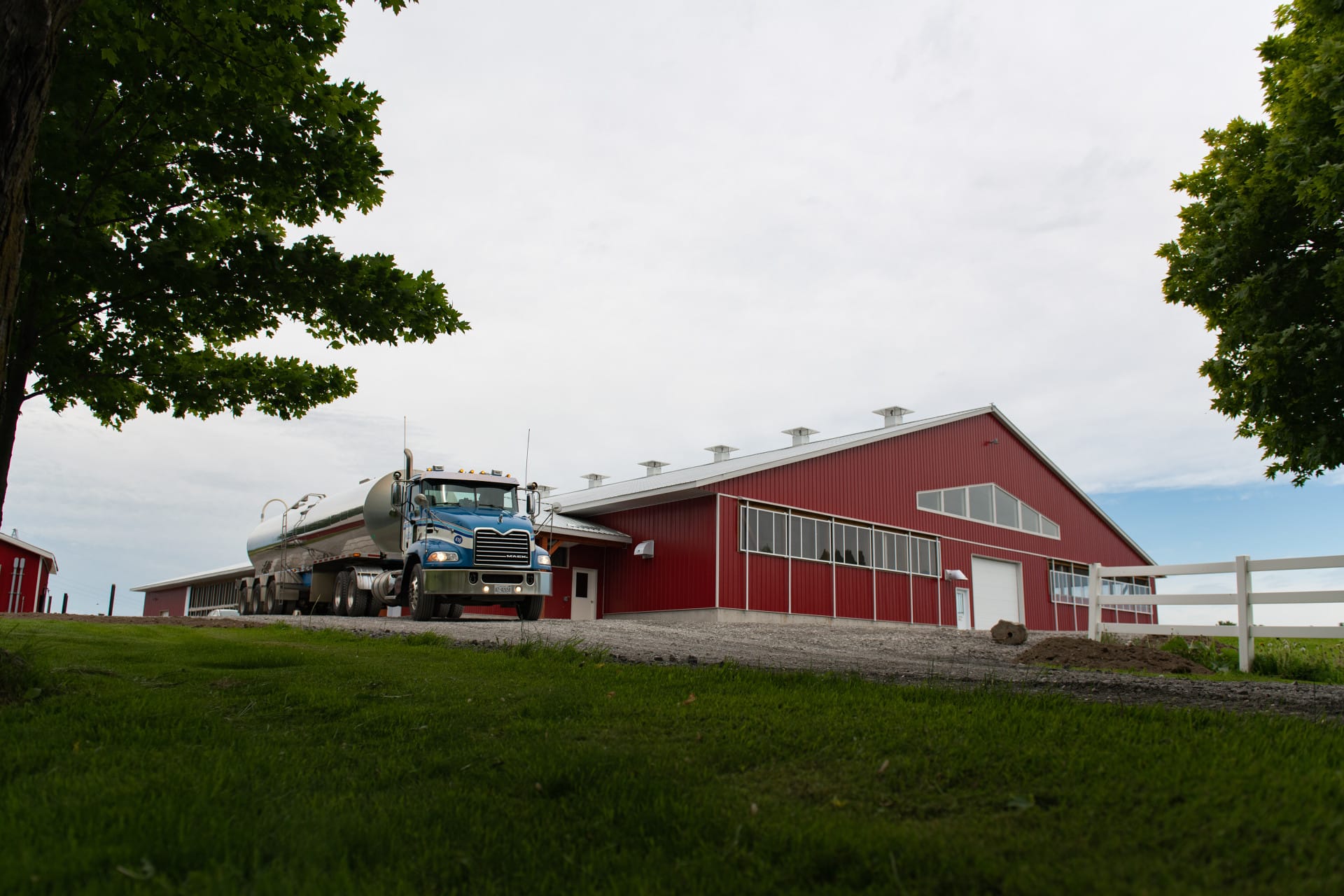 A modern red barn with large windows and a parked milk tanker truck, set against a backdrop of green grass and an overcast sky, representing a well-maintained agricultural facility.