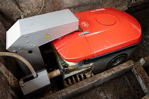 Red circular saw with a metal housing and a warning label on the silver guard, situated above a conveyor or track for materials to be cut. This industrial machinery is used for cutting materials in manufacturing or construction.