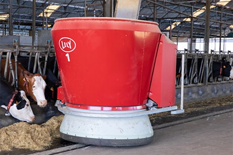 Red, circular robotic device labeled ‘Lely 1’ in a dairy farm setting, situated on a bed of straw with cows in the background. This device appears to be a robotic feed pusher, showcasing modern agricultural technology used to automate livestock feeding processes.