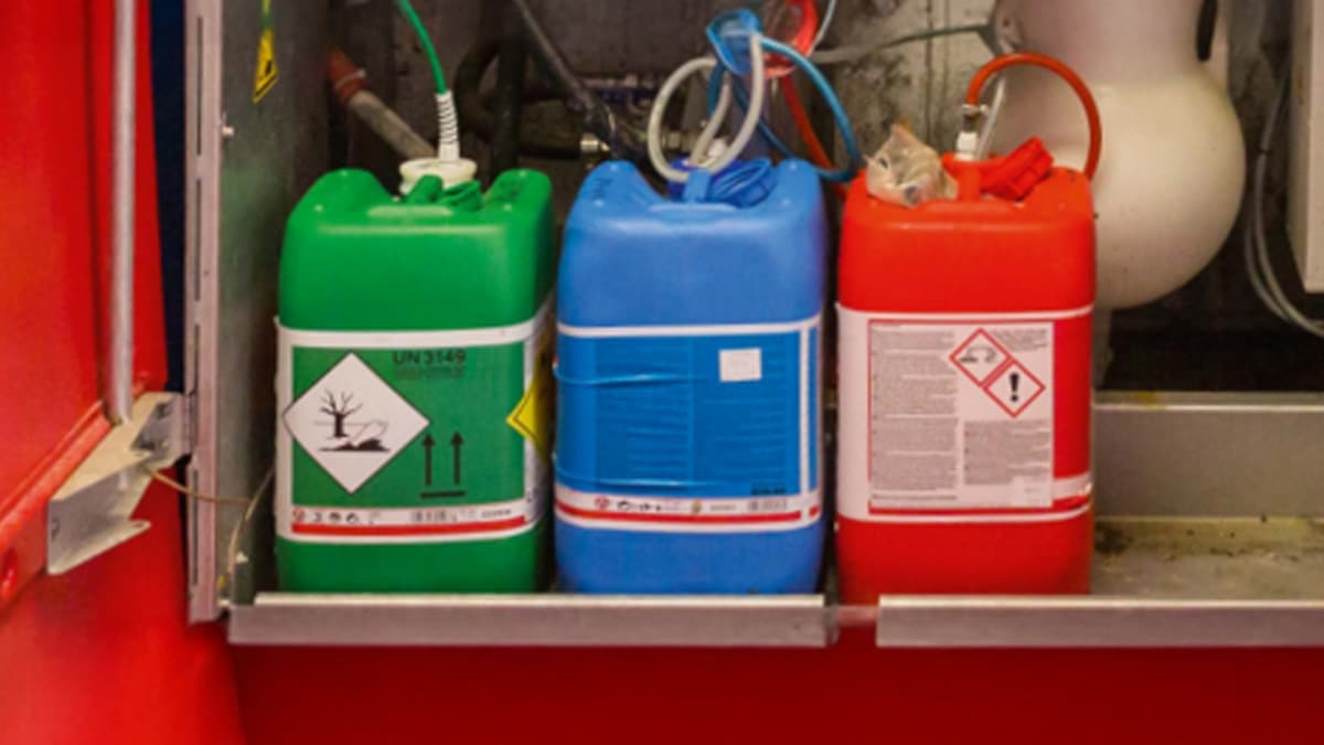 Three industrial containers with hazard symbols stored on a shelf within a red compartment. The green container has a flammable symbol, the blue container appears to be for chemical storage, and the red container has an oxidizing agent symbol.