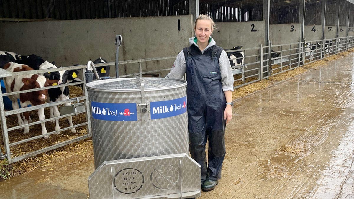 Individual in a blue overall standing next to a large metallic milk container labeled ‘Milk Taxi’ in a cattle barn, with cows visible in the background. The ground is muddy, and the structure behind has numbers 1 through 6, indicating different sections or stalls.